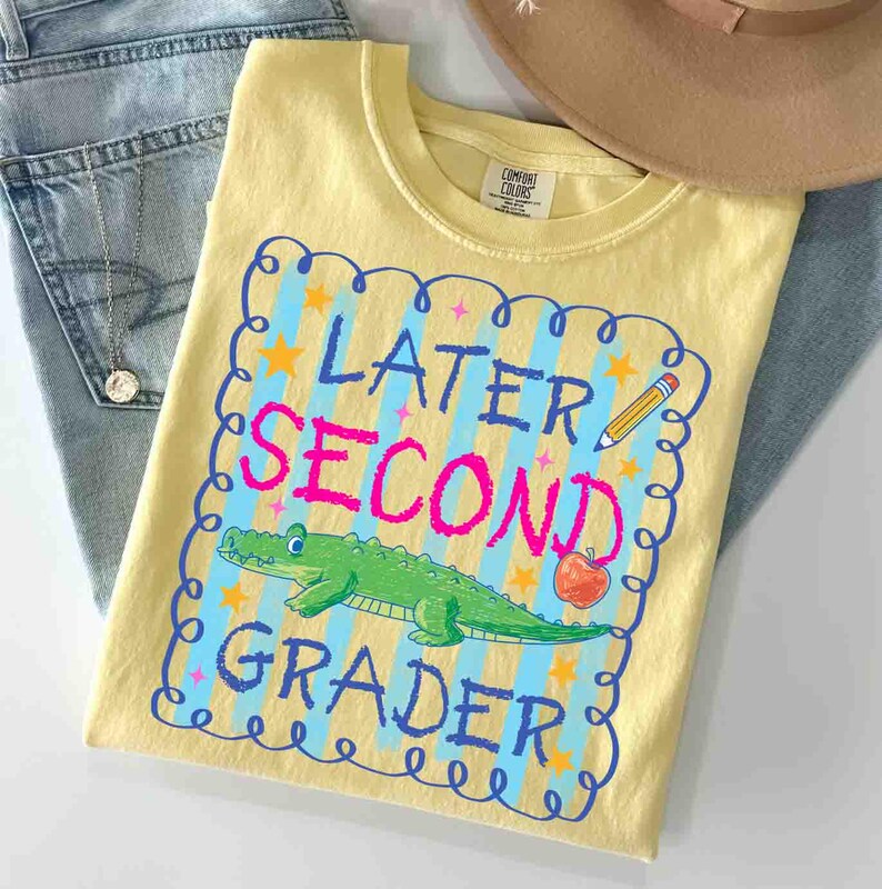 Op de afbeelding: Een geel t-shirt met de woorden "Later Second Grader" in blauw en roze, met een groene alligator en een appel. Het shirt ligt op een blauwe spijkerbroek en in de buurt van een bruine hoed.