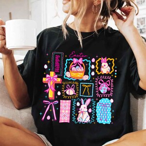 May include: Black t-shirt with a colorful Easter-themed graphic. The design includes a cross, Easter eggs, bunnies, and bows, with the word "Happy" and "Easter" in the design. The graphic is framed with decorative borders.
