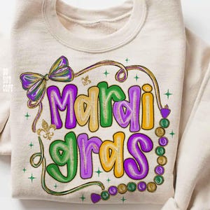May include: A beige sweatshirt with a colorful "Mardi Gras" graphic. The design features purple, green, and yellow lettering, a bow, and a strand of beads. Gold glitter accents and star details complete the festive look.