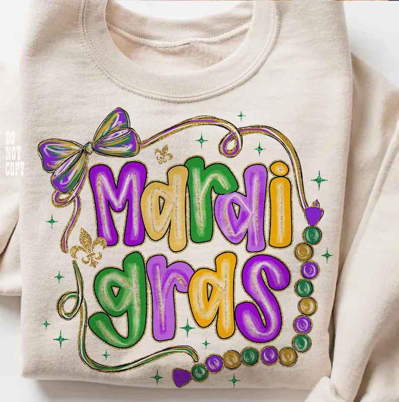 May include: A beige sweatshirt with a colorful "Mardi Gras" graphic. The design features purple, green, and yellow lettering, a bow, and a strand of beads. Gold glitter accents and star details complete the festive look.