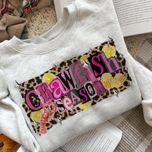 May include: A light gray sweatshirt with a graphic that reads "Crawfish Season" in pink and black letters. The graphic includes a crawfish, lemon slices, and star accents on a leopard print background.