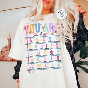 May include: White t-shirt with a colorful alphabet chart that says "You Are" at the top. Each letter of the alphabet is paired with a positive adjective. For example, "A" is for "Accepting" and "B" is for "Brave".
