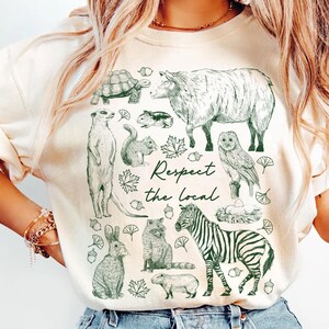 May include: Cream-colored sweatshirt featuring a green illustration of various animals, including a turtle, sheep, meerkat, squirrel, owl, rabbit, fox, zebra, and capybara. The text "Respect the local" is also printed on the shirt.