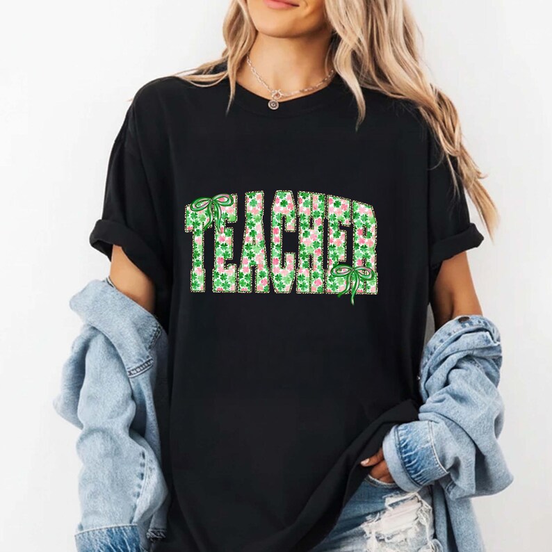 May include: Black t-shirt with the word "TEACHER" in a green and pink shamrock pattern. The word is outlined in green and has green bows on the top and bottom. The shirt is a casual style.