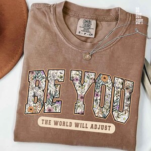 May include: A brown t-shirt with the words "BE YOU" in a floral design. Below the text is the phrase "THE WORLD WILL ADJUST". The shirt has a Comfort Colors tag and a silver necklace.
