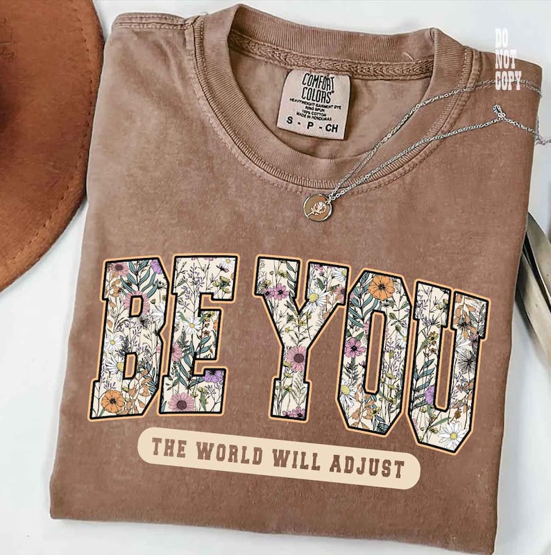 May include: A brown t-shirt with the words "BE YOU" in a floral design. Below the text is the phrase "THE WORLD WILL ADJUST". The shirt has a Comfort Colors tag and a silver necklace.