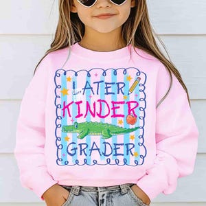 Op de afbeelding: Een lichtroze sweatshirt met de tekst "Later Kinder Grader" in een blauw en roze kader. Het ontwerp bevat een groene alligator, een rode appel, een potlood en sterren.