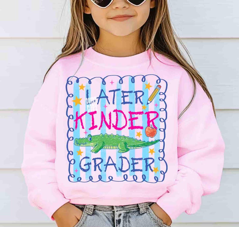 Op de afbeelding: Een lichtroze sweatshirt met de tekst "Later Kinder Grader" in een blauw en roze kader. Het ontwerp bevat een groene alligator, een rode appel, een potlood en sterren.