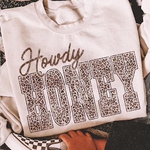 May include: Cream-colored sweatshirt with the words "Howdy HONEY" printed on the front. The word "Howdy" is in a cursive font, and "HONEY" is in a block letter style with a leopard print pattern. The sweatshirt is casual wear.