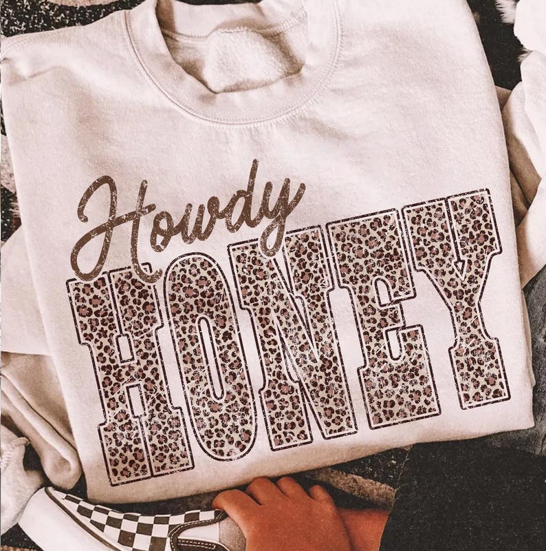 May include: Cream-colored sweatshirt with the words "Howdy HONEY" printed on the front. The word "Howdy" is in a cursive font, and "HONEY" is in a block letter style with a leopard print pattern. The sweatshirt is casual wear.