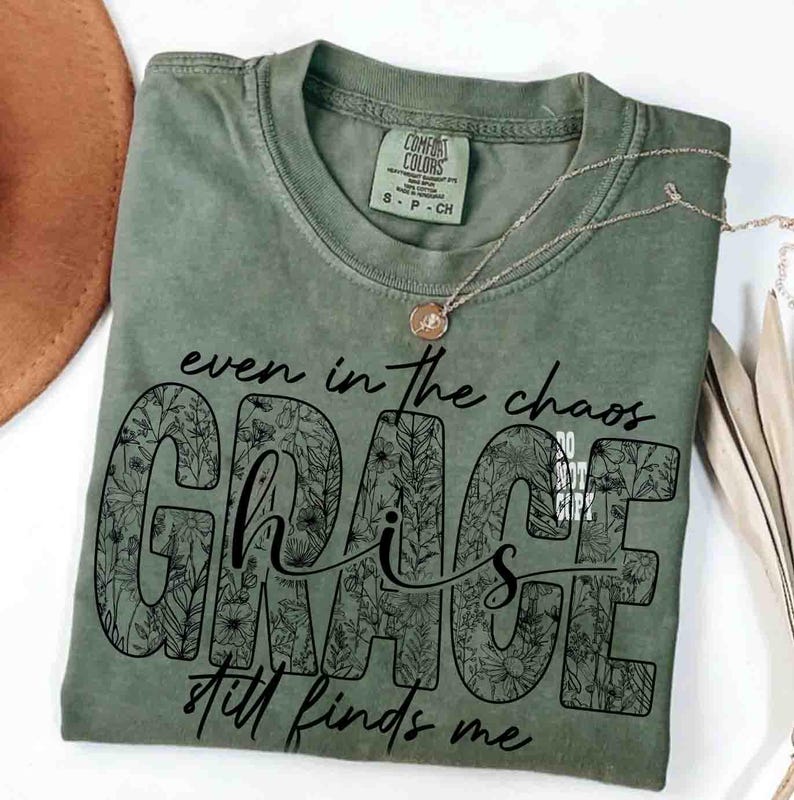 May include: An olive green Comfort Colors t-shirt with the words "even in the chaos GRACE still finds me" in black script. The word "GRACE" is filled with a floral design. A gold necklace with a pendant rests on the shirt.