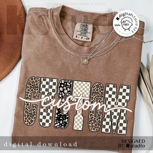 Może przedstawiać: Brązowa koszulka Comfort Colors z napisem "CUSTOM" w odważnym, wzorzystym designie. Litery mają wzory w cętki, szachownicę i grochy. Srebrny naszyjnik z wisiorkiem jest zarzucony na koszulkę. Słowa "digital download" znajdują się na dole.