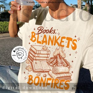Könnte beinhalten: Cremefarbenes T-Shirt mit orangefarbener Grafik, die gestapelte Bücher, ein Lagerfeuer, gerollte Decken und die Worte "Books, Blankets, Bonfires" zeigt. Das Design enthält Herbstblätter und Sterne. "Digital download" ist unten aufgedruckt.