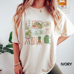 May include: Ivory-colored t-shirt featuring a colorful Easter-themed graphic. The design includes a bunny, eggs, carrots, and other spring elements arranged in a grid pattern. The shirt has short sleeves and a relaxed fit.