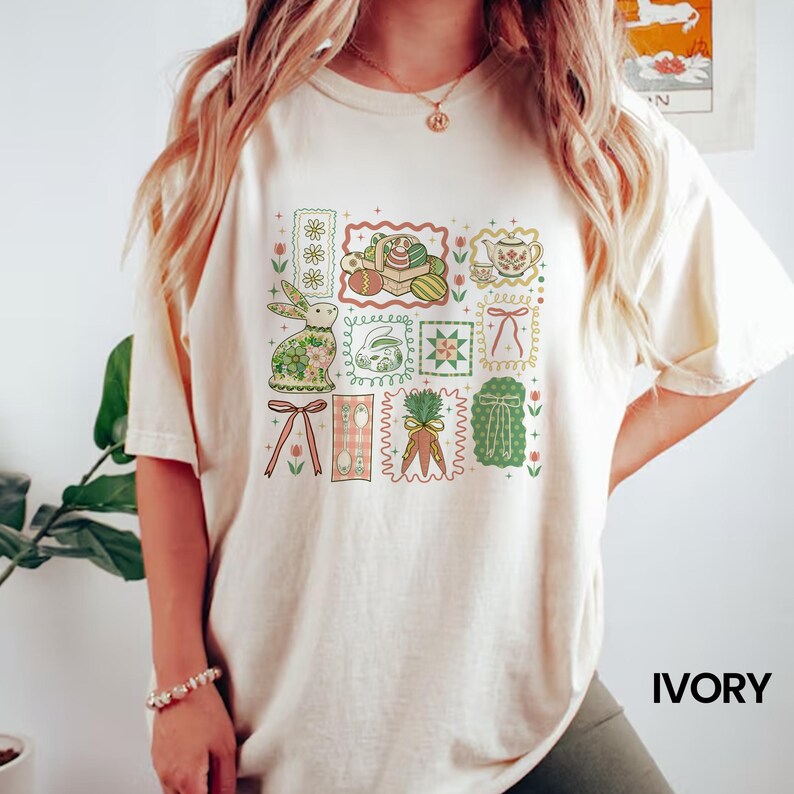 May include: Ivory-colored t-shirt featuring a colorful Easter-themed graphic. The design includes a bunny, eggs, carrots, and other spring elements arranged in a grid pattern. The shirt has short sleeves and a relaxed fit.