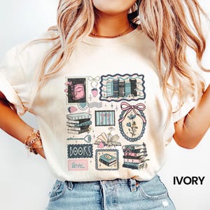 May include: Ivory-colored t-shirt featuring a collage of book-themed illustrations. The design includes stacks of books, open books, and floral elements in pink, blue, and green. The word "BOOKS" and "love" are also included in the design.