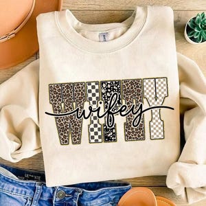 May include: A cream-coloured sweatshirt featuring the word "wifey" in a decorative font. The letters are filled with patterns like leopard print, checkered, and polka dots. A black cursive font overlays the word "wifey".
