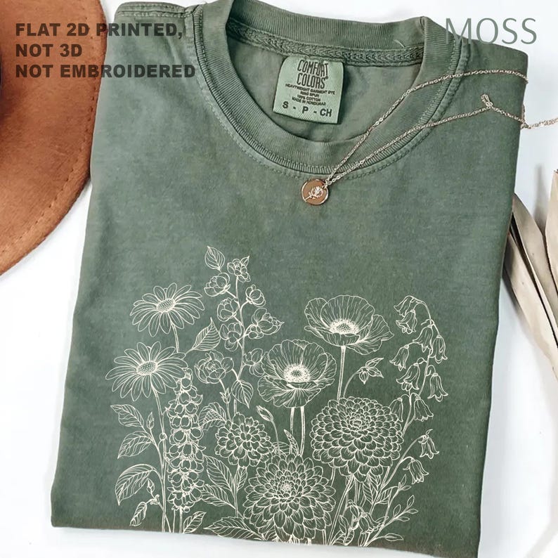 May include: A moss green t-shirt with a white floral design. The shirt has a crew neck and short sleeves. A silver necklace with a small pendant rests on the shirt. The text "FLAT 2D PRINTED, NOT 3D, NOT EMBROIDERED" is visible.