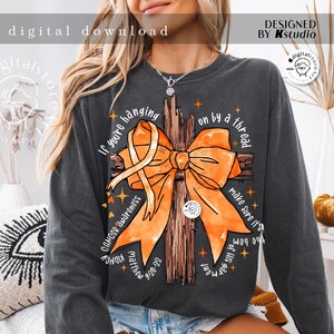 May include: A grey long-sleeved shirt with an orange ribbon tied in a bow around a wooden cross. The text "If you're hanging on by a thread make sure it's the hem of His garment" is printed on the shirt.  Cancer awareness, Matthew 9:20-22, and KIDNE are also printed on the shirt.