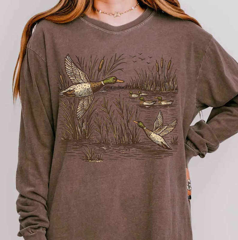 May include: A brown long-sleeve shirt featuring a nature-themed graphic. The graphic depicts ducks flying over a pond with reeds. The ducks have green heads and brown wings. The shirt is a comfortable, casual style.