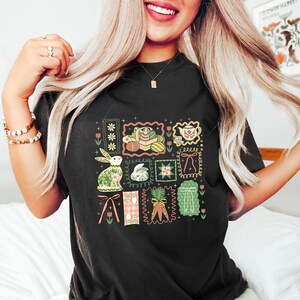 May include: Black t-shirt featuring a colorful Easter-themed graphic. The design includes framed illustrations of Easter eggs, bunnies, carrots, and floral elements. The artwork is in a vintage, patchwork style, with pastel colors and decorative borders.