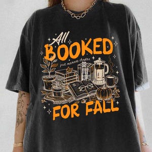 May include: Black t-shirt with an orange graphic that reads "All Booked For Fall." The design includes illustrations of books, a pumpkin, coffee, and plants, evoking a cozy autumn theme.
