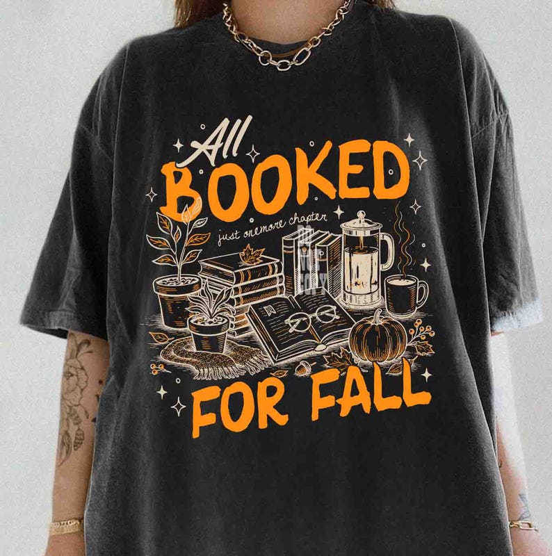 May include: Black t-shirt with an orange graphic that reads "All Booked For Fall." The design includes illustrations of books, a pumpkin, coffee, and plants, evoking a cozy autumn theme.