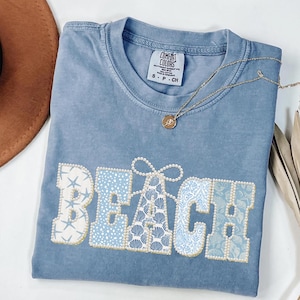 May include: A blue Comfort Colors t-shirt with the word "BEACH" in a decorative font. The letters are embellished with seashells, starfish, and pearls. A gold necklace with a pendant is draped across the shirt.