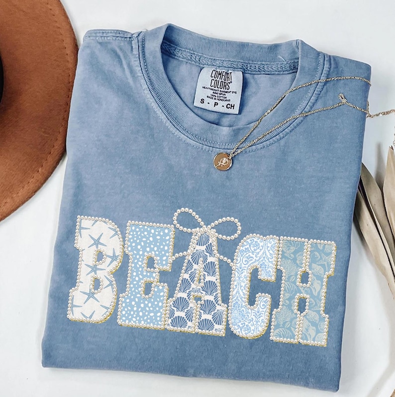 May include: A blue Comfort Colors t-shirt with the word "BEACH" in a decorative font. The letters are embellished with seashells, starfish, and pearls. A gold necklace with a pendant is draped across the shirt.