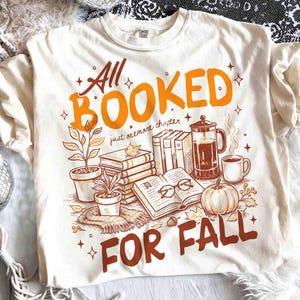 May include: Cream-colored t-shirt with a fall-themed graphic. The design features the text "All Booked For Fall" in orange and brown, along with illustrations of books, a pumpkin, coffee, and plants. The shirt is made of a soft material.