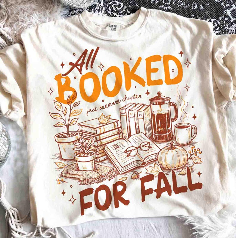 May include: Cream-colored t-shirt with a fall-themed graphic. The design features the text "All Booked For Fall" in orange and brown, along with illustrations of books, a pumpkin, coffee, and plants. The shirt is made of a soft material.