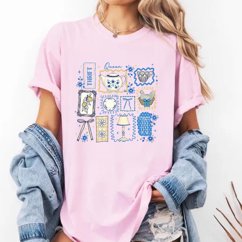 May include: Pink t-shirt featuring a collage of vintage-style illustrations. The design includes a teapot, a plate, a lamp, and other motifs, all in a blue and white color scheme. The word "Thrift" is printed vertically on the left side of the shirt.