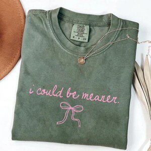 May include: Olive green t-shirt with the phrase "i could be meaner" in pink script and a pink bow graphic. A gold necklace with a pendant is on the shirt. A brown hat is partially visible in the background.