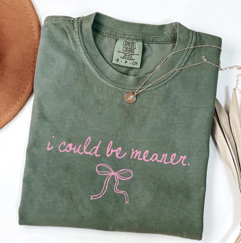 May include: Olive green t-shirt with the phrase "i could be meaner" in pink script and a pink bow graphic. A gold necklace with a pendant is on the shirt. A brown hat is partially visible in the background.