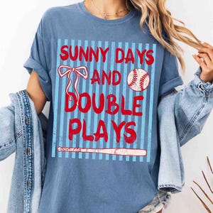 May include: Blue t-shirt with the text "SUNNY DAYS AND DOUBLE PLAYS" in red. The design includes a baseball, a bow, and a baseball bat. The shirt has rolled-up sleeves and a casual fit.