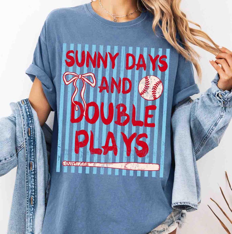 May include: Blue t-shirt with the text "SUNNY DAYS AND DOUBLE PLAYS" in red. The design includes a baseball, a bow, and a baseball bat. The shirt has rolled-up sleeves and a casual fit.