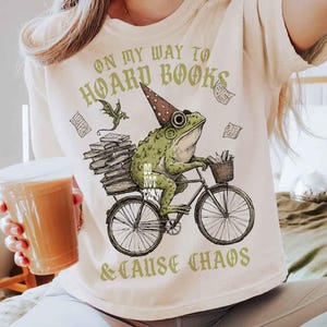 May include: A cream-coloured t-shirt featuring a green frog in a wizard hat riding a bicycle, carrying books. The text reads "ON MY WAY TO HOARD BOOKS & CAUSE CHAOS". The design includes a dragon and floating papers. A fun, book-themed design.