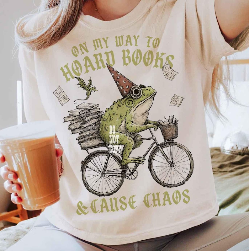 May include: A cream-coloured t-shirt featuring a green frog in a wizard hat riding a bicycle, carrying books. The text reads "ON MY WAY TO HOARD BOOKS & CAUSE CHAOS". The design includes a dragon and floating papers. A fun, book-themed design.
