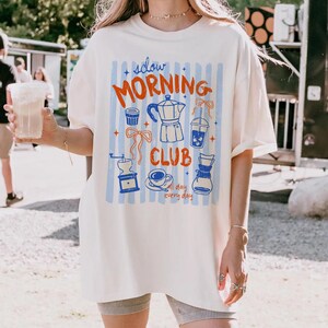 May include: Cream-colored t-shirt with a "Slow Morning Club" graphic in red and blue. The design includes coffee-related illustrations like a coffee maker, cup, and grinder, set against blue vertical stripes. The shirt is a relaxed fit.