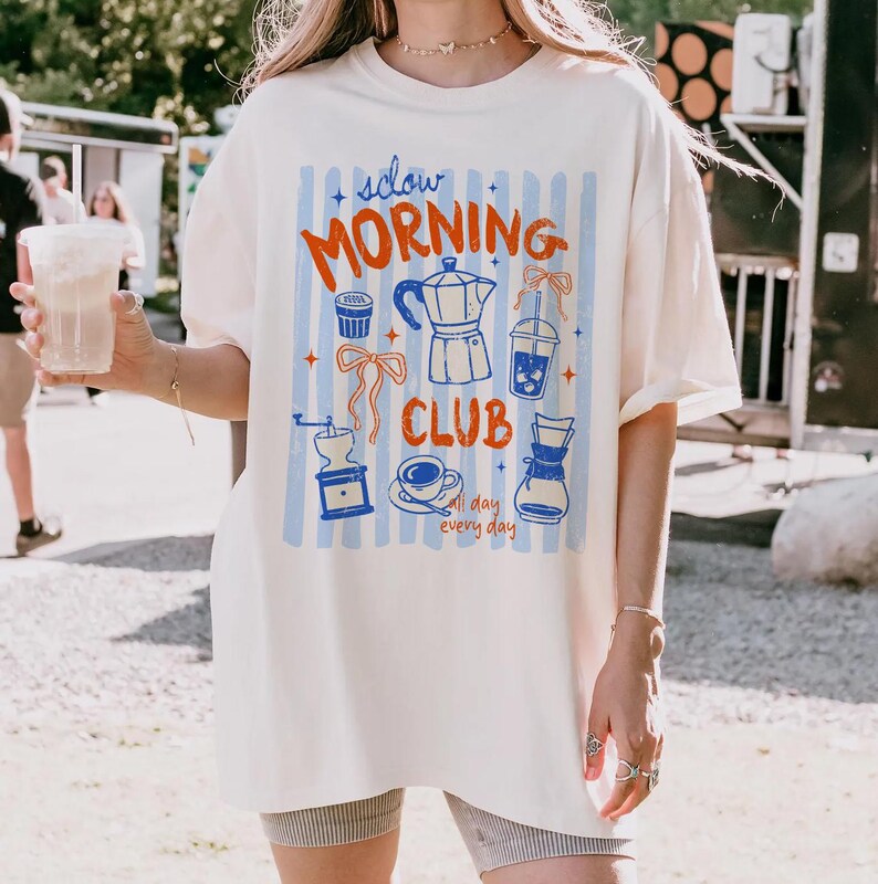 May include: Cream-colored t-shirt with a "Slow Morning Club" graphic in red and blue. The design includes coffee-related illustrations like a coffee maker, cup, and grinder, set against blue vertical stripes. The shirt is a relaxed fit.