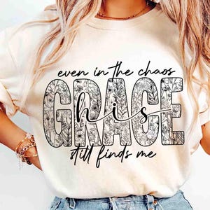May include: Cream-colored t-shirt with the words "even in the chaos, His Grace still finds me" in black script. The word "GRACE" is filled with a floral pattern.