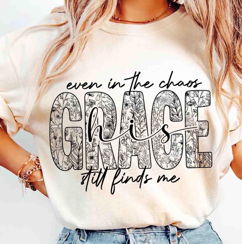May include: Cream-colored t-shirt with the words "even in the chaos, His Grace still finds me" in black script. The word "GRACE" is filled with a floral pattern.