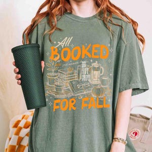 May include: Sage green t-shirt with the text "All Booked For Fall" in orange. The graphic includes illustrations of books, plants, a pumpkin, and a coffee pot. A dark green tumbler is held in the hand.