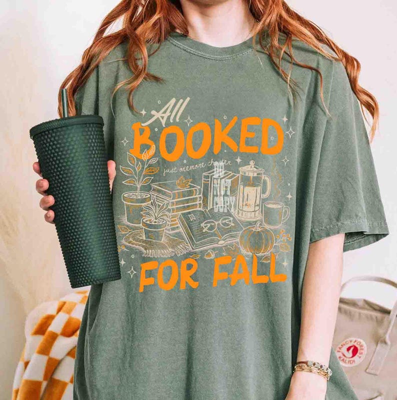 May include: Sage green t-shirt with the text "All Booked For Fall" in orange. The graphic includes illustrations of books, plants, a pumpkin, and a coffee pot. A dark green tumbler is held in the hand.