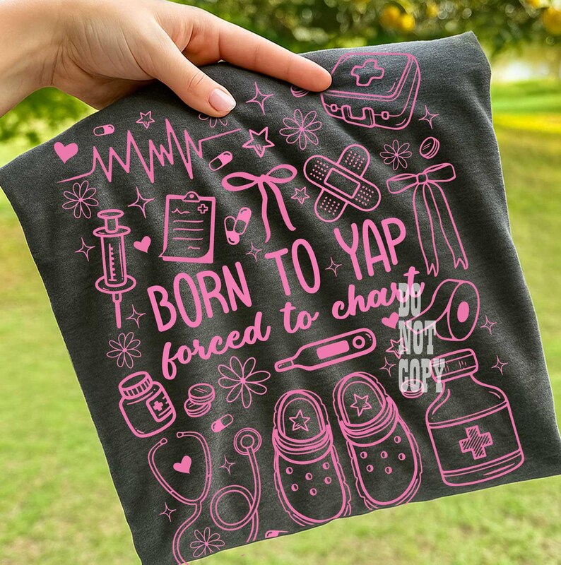 K&ouml;nnte beinhalten: Dunkelgraues T-Shirt mit rosa medizinischen Grafiken, darunter ein Stethoskop, eine Spritze und eine Medikamentenflasche. Der Text "BORN TO YAP forced to chart" ist in Rosa dargestellt. Das Shirt zeigt auch Clogs und einen Erste-Hilfe-Kasten.