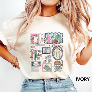 Op de afbeelding: Ivoor kleurig T-shirt met een collage van medische illustraties in postzegelachtige kaders. Het ontwerp bevat een stethoscoop, een spuit, pillen en een koffiekopje. Het woord "Nurse" is ook zichtbaar.