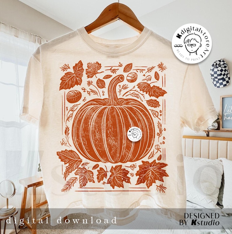 以下が含まれることがあります： オレンジと茶色のグラフィックデザインが施されたベージュのTシャツ。デザインは、葉やその他の秋の要素に囲まれたカボチャです。画像の右上に「Kdigitalstoreart」というテキストが表示されています。