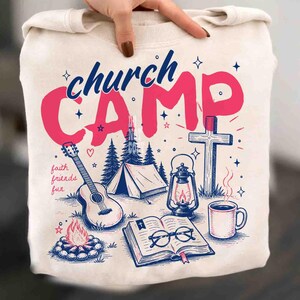 K&ouml;nnte beinhalten: Cremefarbenes Sweatshirt mit einem grafischen Design mit den Worten "church CAMP" in Pink und Blau. Das Design umfasst eine Gitarre, ein Zelt, ein Lagerfeuer, ein Kreuz, eine Laterne, eine Kaffeetasse und ein offenes Buch mit Brille. Die Worte "faith friends fun" sind ebenfalls enthalten.