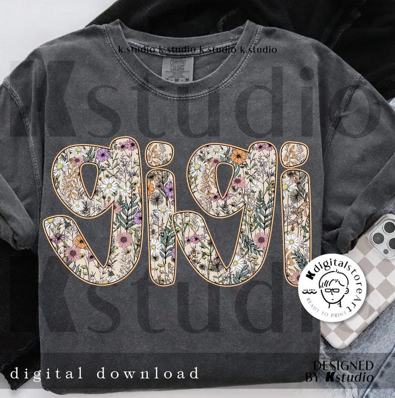 May include: A gray t-shirt with the word "gigi" in large, white letters outlined in orange. The letters are filled with a floral pattern of pink, purple, and yellow flowers.