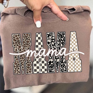 May include: A brown sweatshirt with the word "MAMA" in large, decorative letters. The letters have various patterns, including leopard print, checkered, and speckled designs. The word "mama" is written in cursive below.